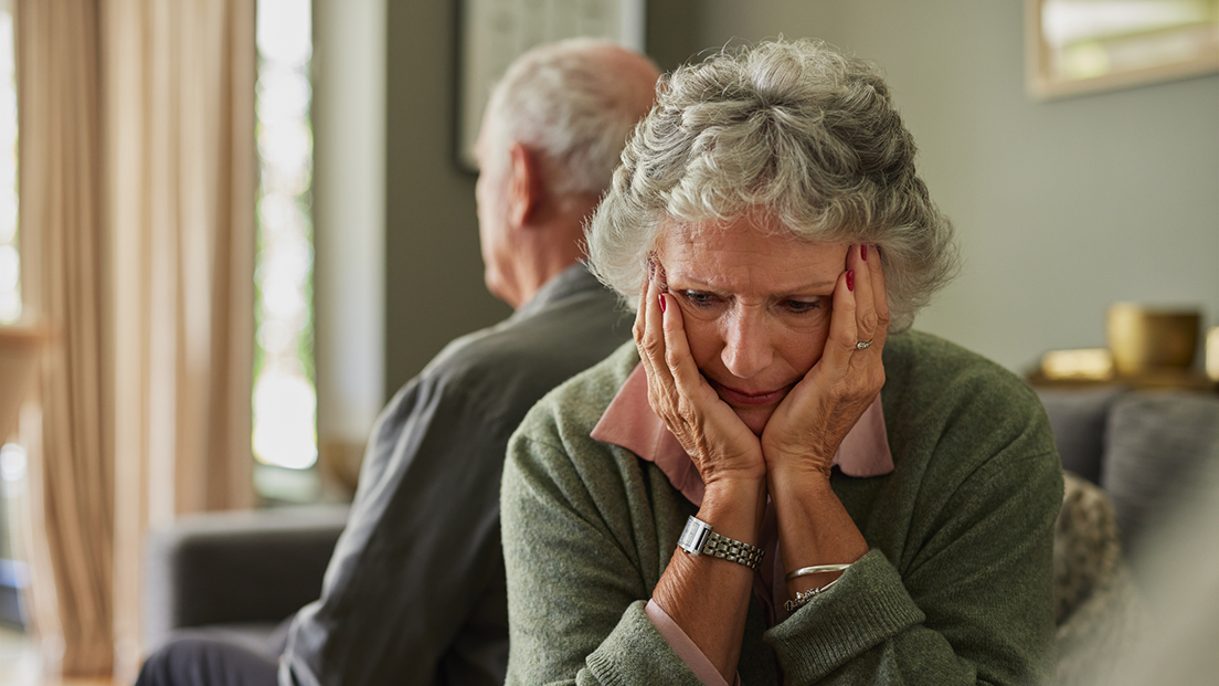 An older woman sits on a couch with her head in her hands, looking sad and deep in thought. Behind her, an older man sits turned away, also appearing distant. The mood is tense and somber, suggesting emotional strain or disconnection between them in a quiet living room setting.