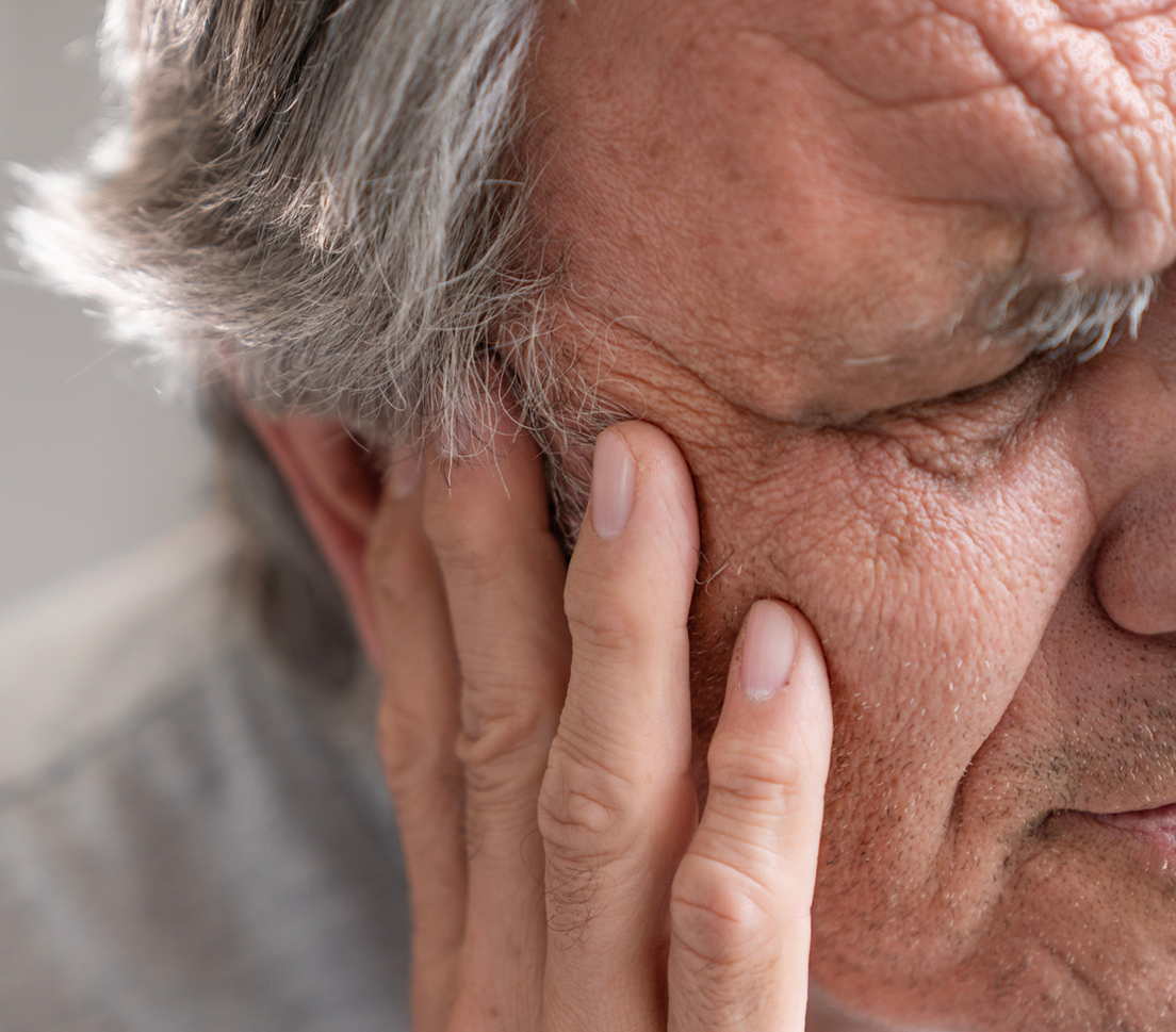 Close-up of an older man touching his ear with two fingers, suggesting ear discomfort or pain.