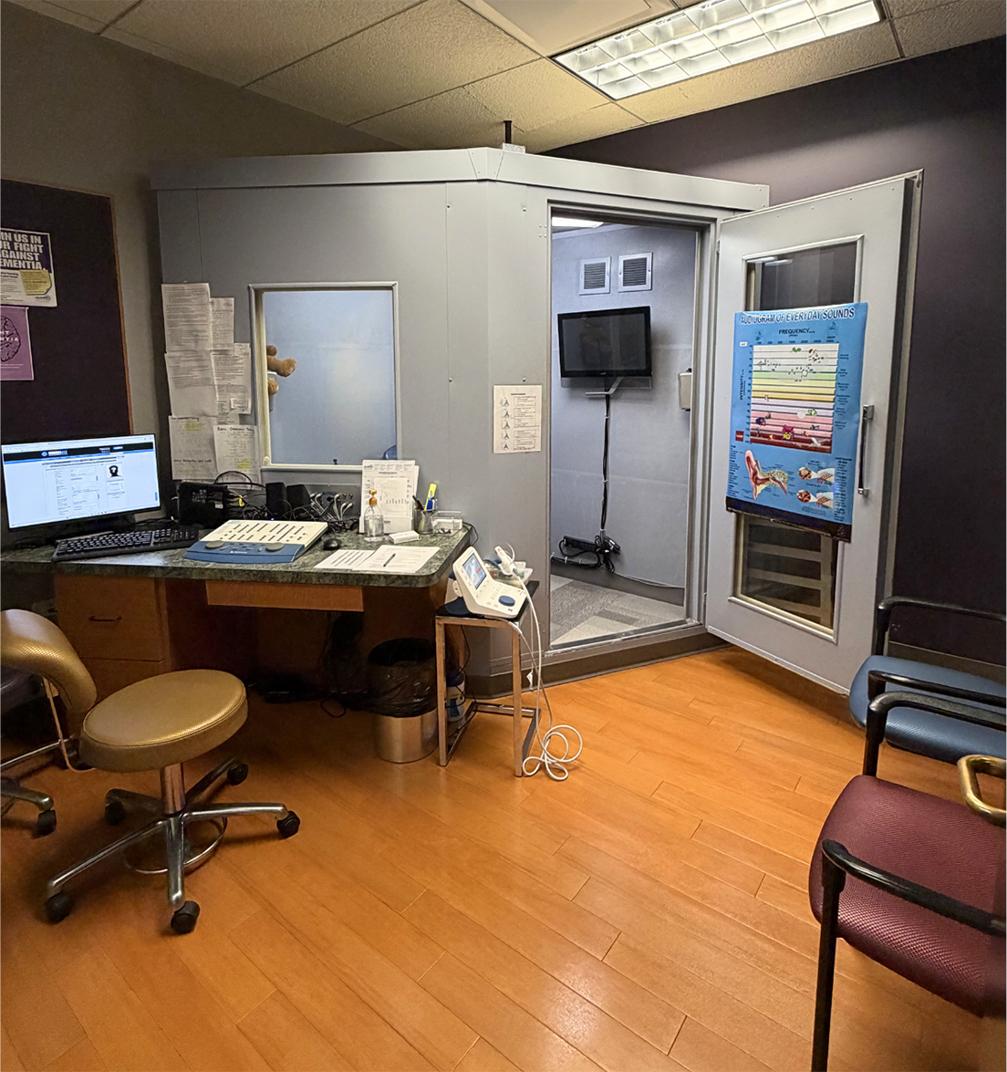 A small clinic with a windowed sound booth door open, next to a desk with a computer and medical equipment.